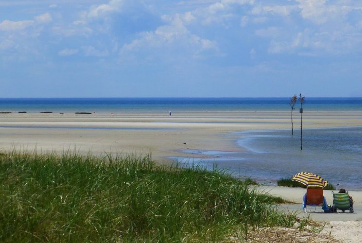 Expansive beach area near bright blue waters with two people sitting in beach chairs near green dune grass