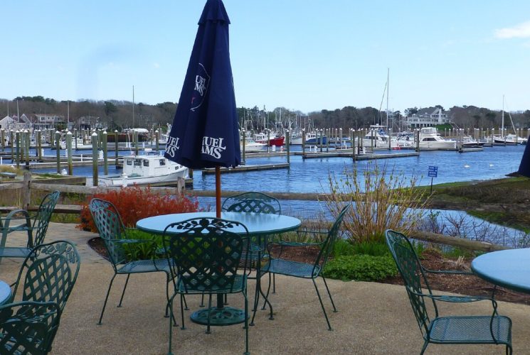 Outdoor patio dining area with green tables and chairs at waters edge with marina in background