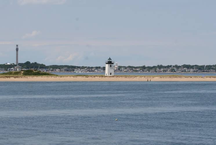 Small black and white lighthouse at the end of a small peninsula surrounded by water with with a city in the background