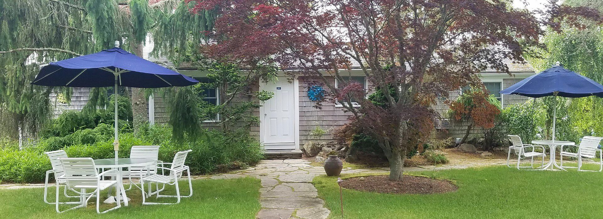 Front facade of a small home with lots of trees, lawn and two patio tables with chairs and blue umbrellas