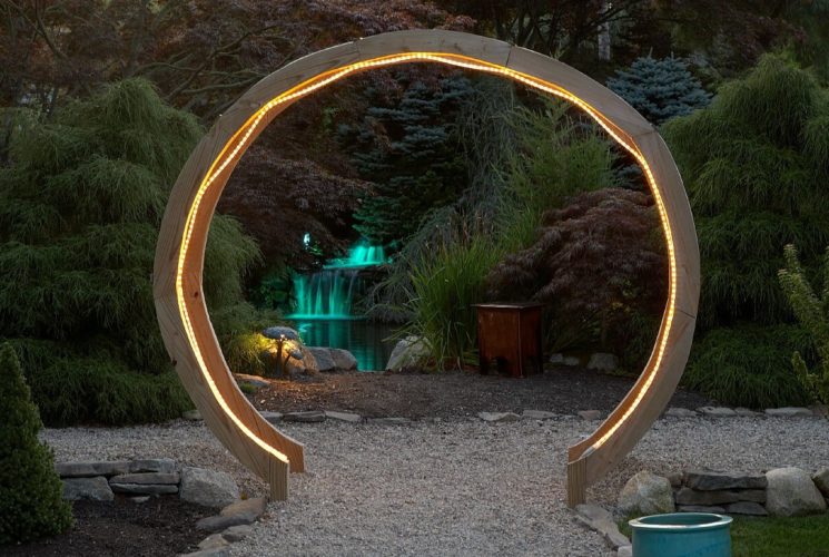 Large circular wooden gate with rope lights lit up at night with colorful green fountain in the background