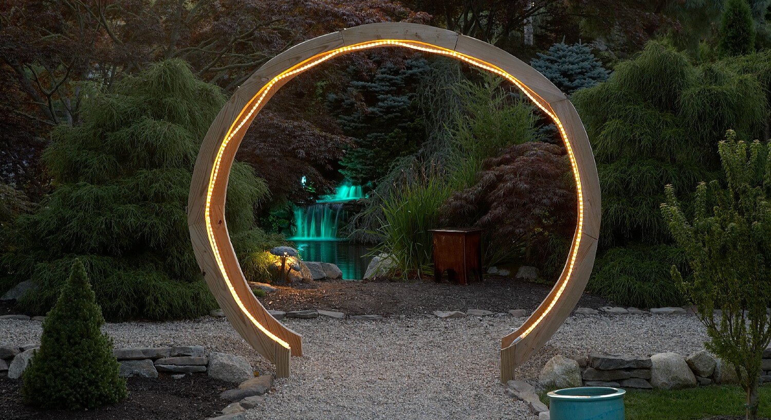 Large circular wooden gate with rope lights lit up at night with colorful green fountain in the background