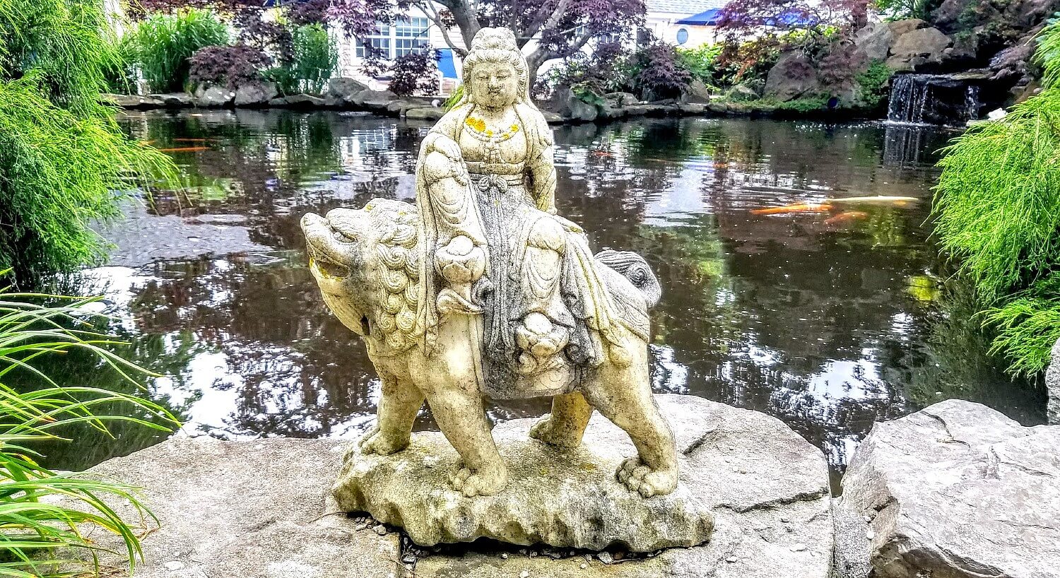 Large stone Asian-inspired statue on top of a large rock at the edge of a pond surrounded by colorful trees and plants
