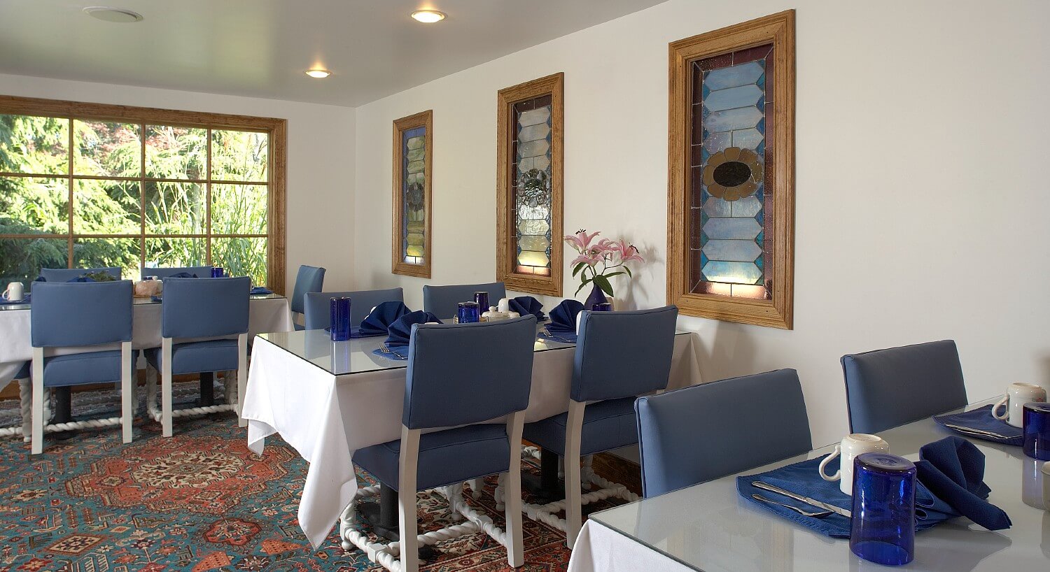 Dining area with stained glass windows and three glass top tables with with blue chairs set for breakfast