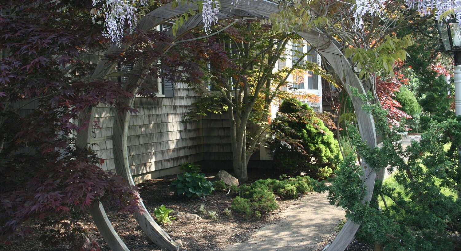 Two wooden arches at the edge of a path leading up to a cottage surrounded by colorful trees and bushes