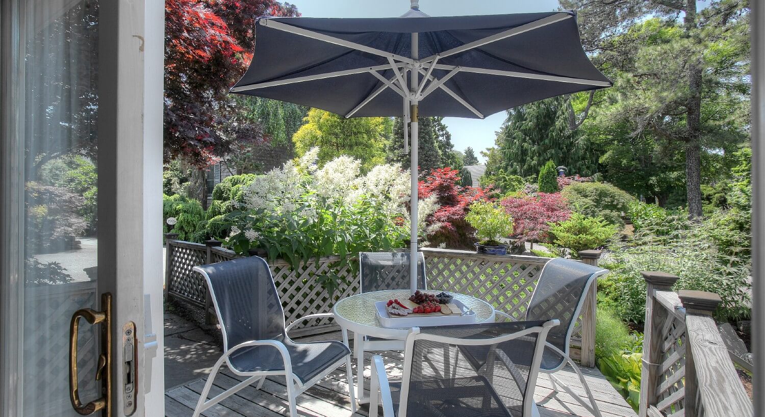 Outdoor deck with patio table, four chairs and umbrella surrounded by lush colorful bushes