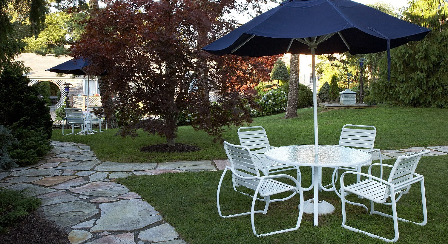 Outdoor lawn area with stone sidewalks and white patio table wth four chairs and blue umbrella