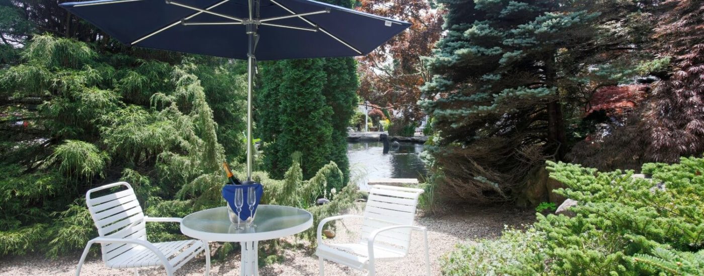 White outdoor patio table with champagne glasses, two chairs and a blue umbrella on stone area surrounded by lush green trees, bushes & Koi pond in the background.