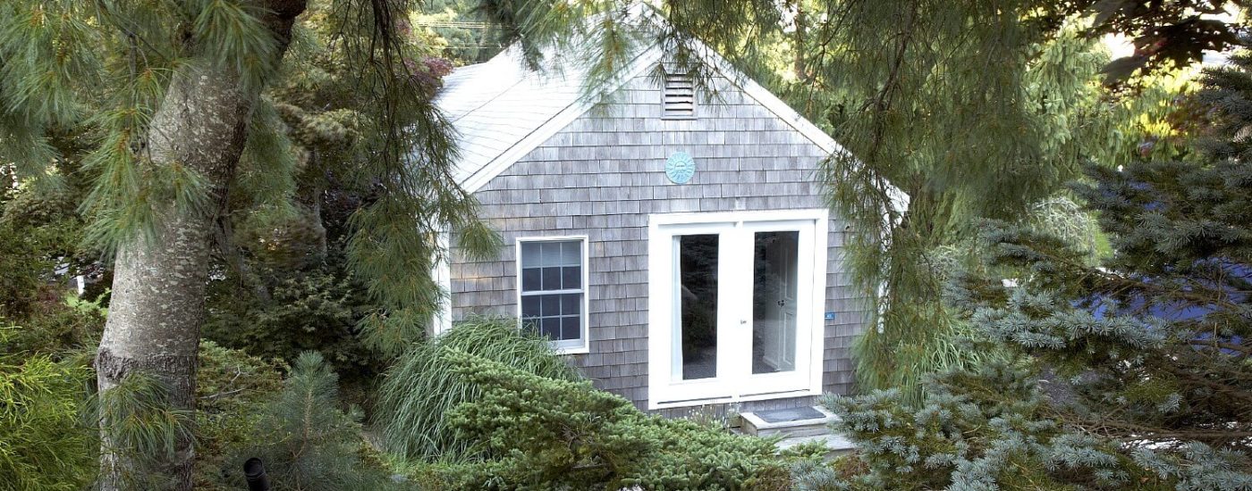 Small cottage with grey shingles and white trim nestled among tall trees and green shrubs