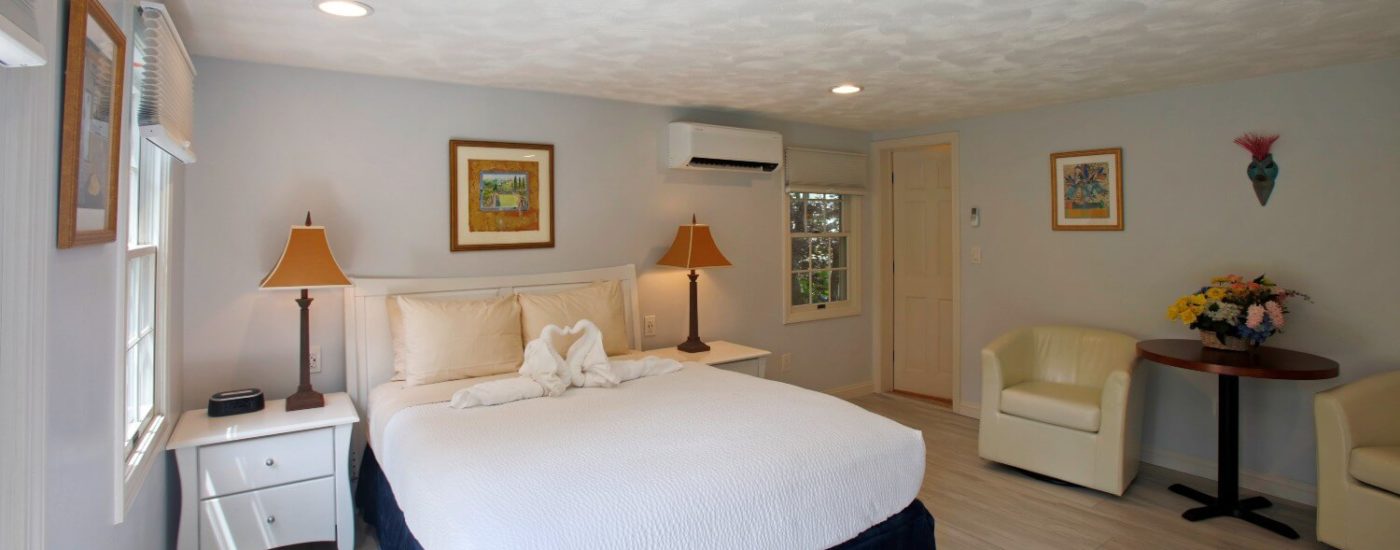 Bright bedroom with queen sized bed in white linens, wood nightstands, white leather chair & light wood flooring.