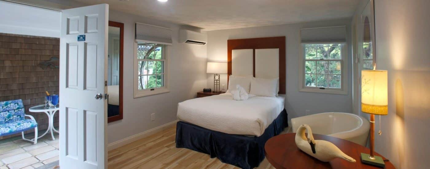 Door from a breezeway open to a bright bedroom with one bed, two windows, sitting chair by a round table and light wood floors.