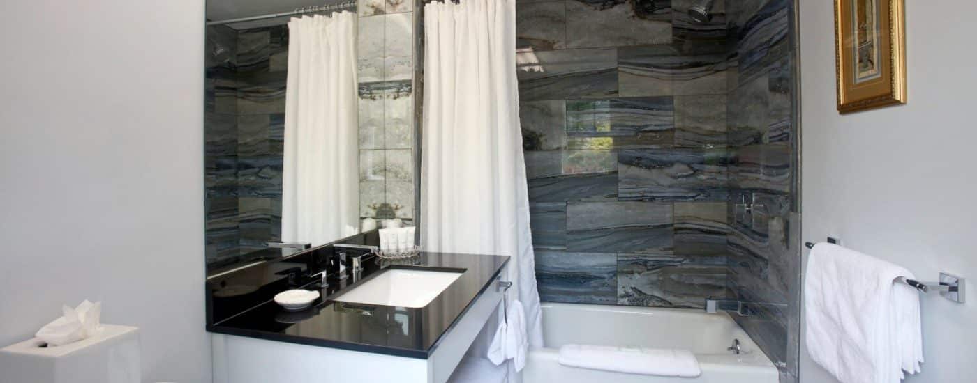 Shiny bathroom with black and grey tiled wall around white bathtub sink with black granite top and large mirror on wall.
