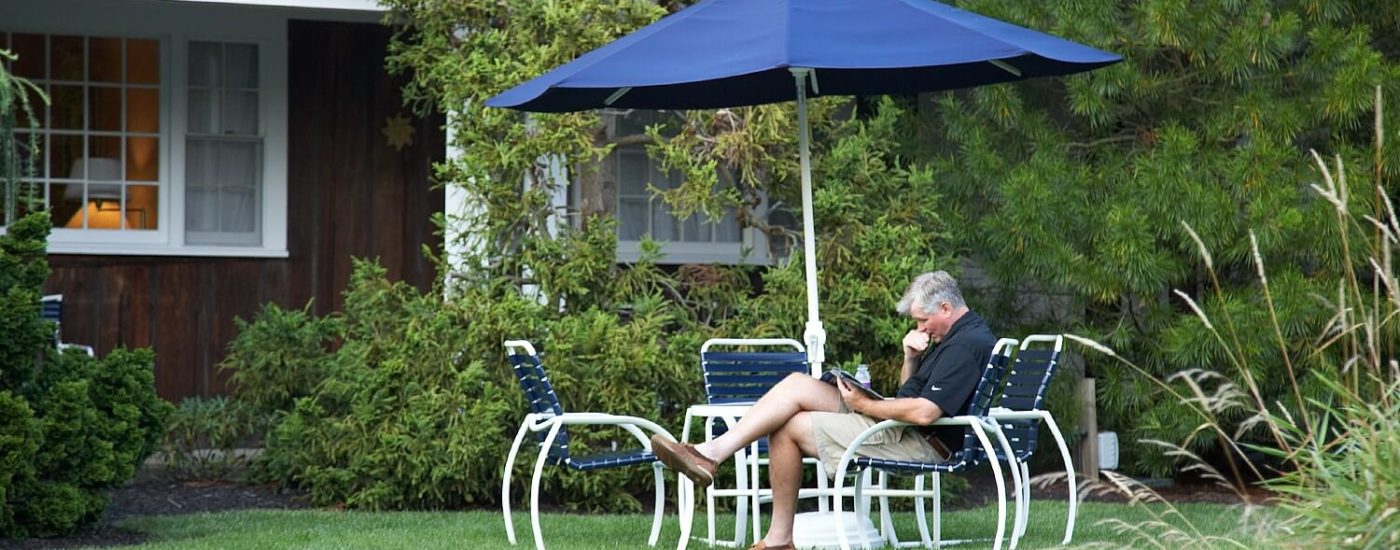 A man in shorts and a black shirt sitting at a white outdoor patio table with chairs and a blue umbrella on a lawn