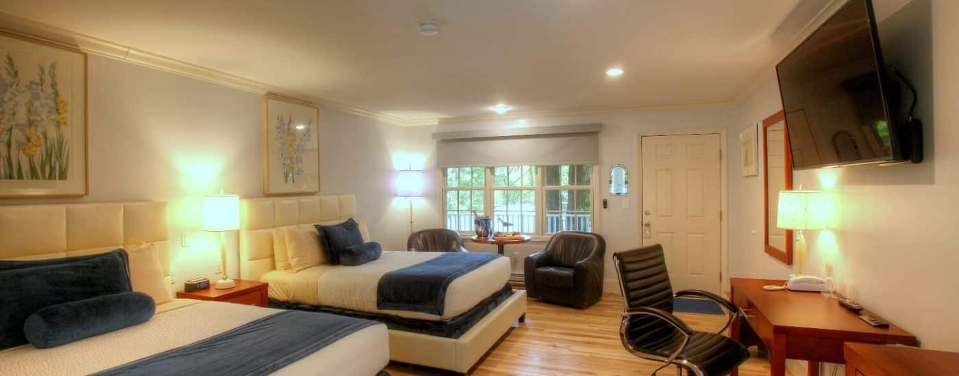 Bedroom with two queen beds in white and blue linens, two leather sitting chairs, and writing table and chair under TV