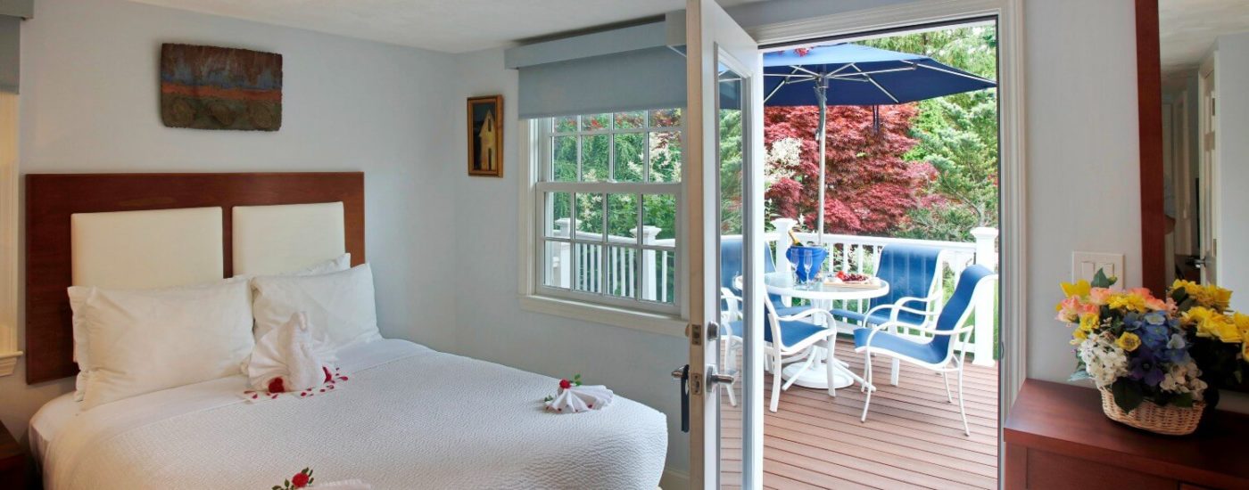 Bright bedroom with queen bed in white linens and door to an outdoor deck with patio furniture & umbrella surrounded by trees.