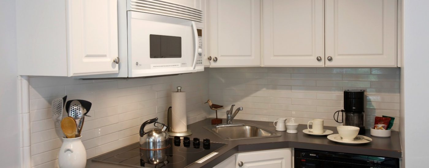 Kitchenette of a studio apartment with black countertop, white cabinets , a cooktop, microwave, sink & dishwasher.