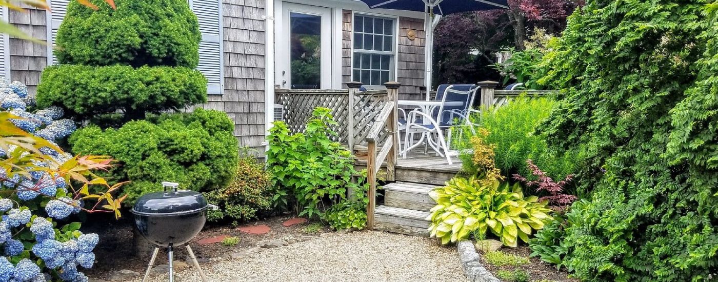 Back deck of a studio apartment with patio table and umbrella, bbq and lush plants and bushes