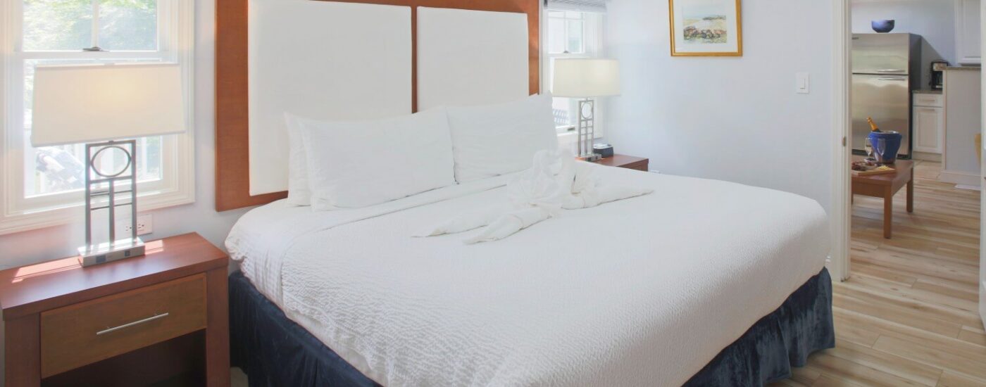 Bedroom with king bed, two bright windows and doorway leading into living area with coffee table and kitchenette