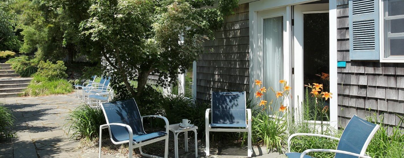 Outdoor patio with lush trees and flowers with blue sitting chairs by an open door leading to a bedroom