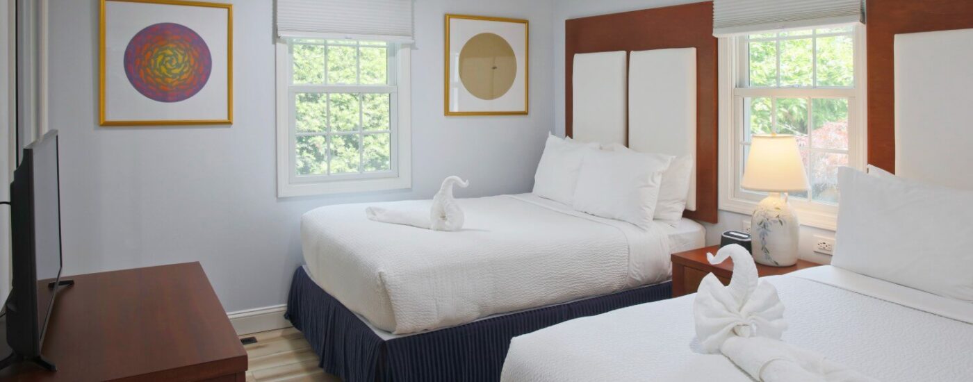 Cottage bedroom with two queen beds in white and blue linens, two windows and dresser with TV