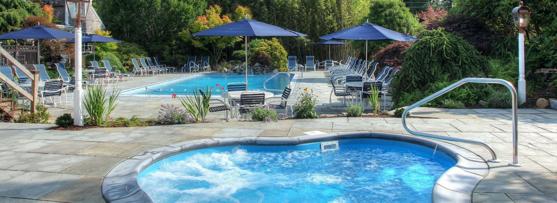 Large outdoor patio with pool lined with sunning chairs, a hot tub with bubbling water and patio tables with blue umbrellas