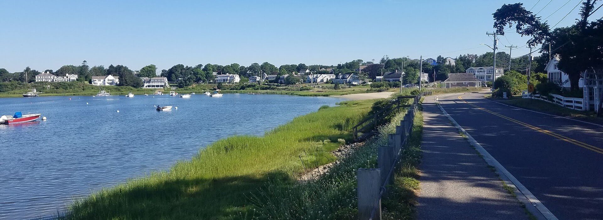 Two lane drive curving next to a body of water with several fishing boats and lined with large homes