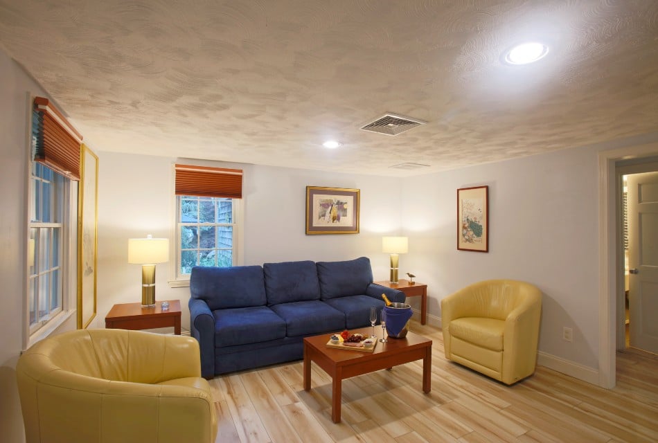 Bright living area of a cottage with light wood floors, two yellow leather chairs, blue sofa, matching wood coffee table & side tables, and light colored wood floor.