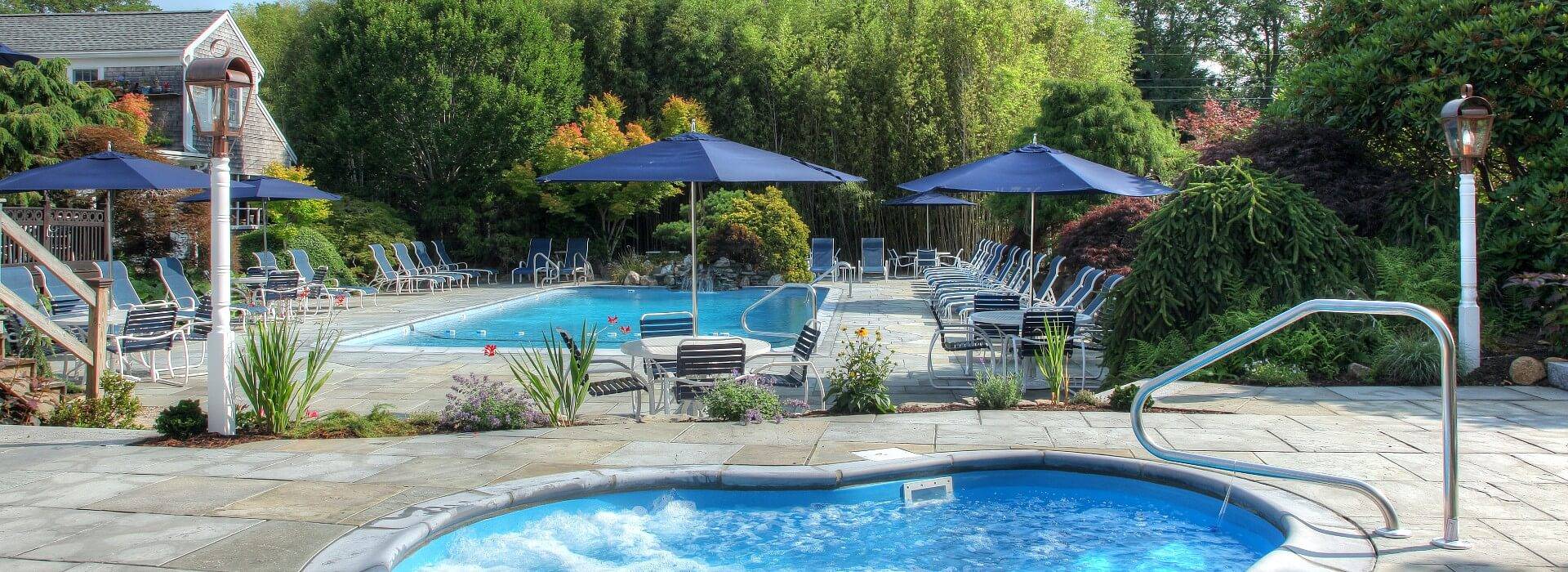 Gorgeous outdoor patio with large pool lined with chairs, bubbling hot tub and patio tables with blue umbrellas