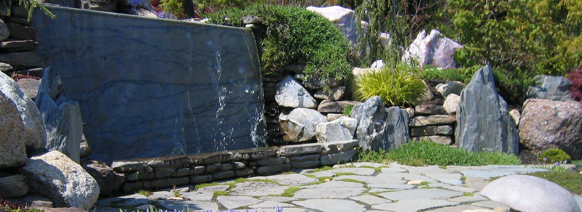 Unique wall waterfall feature surrounded by large rocks, stone patio and green trees and shrubs