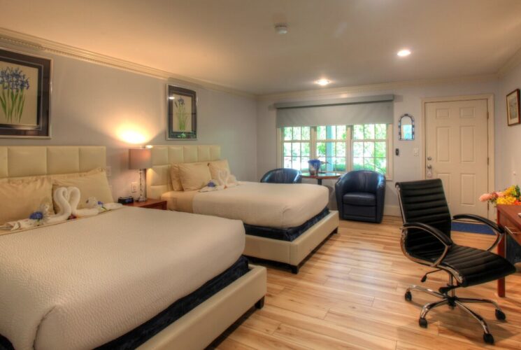 Large guest room with two queen beds, blue leather sitting chair, writing desk underneath TV and arched doorway